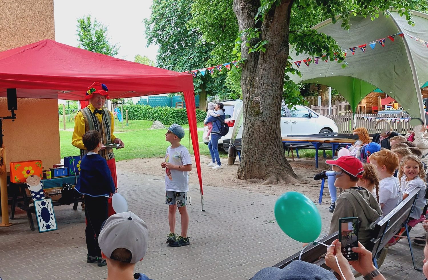 Links steht ein bunter Zauberer der gerade 2 jungen einen Zaubertrick zeigt. Die anderen Schulanfänger sitzen in einem Halbkreis auf Bänken und schauen der Show zu. Es findet im Garten statt.